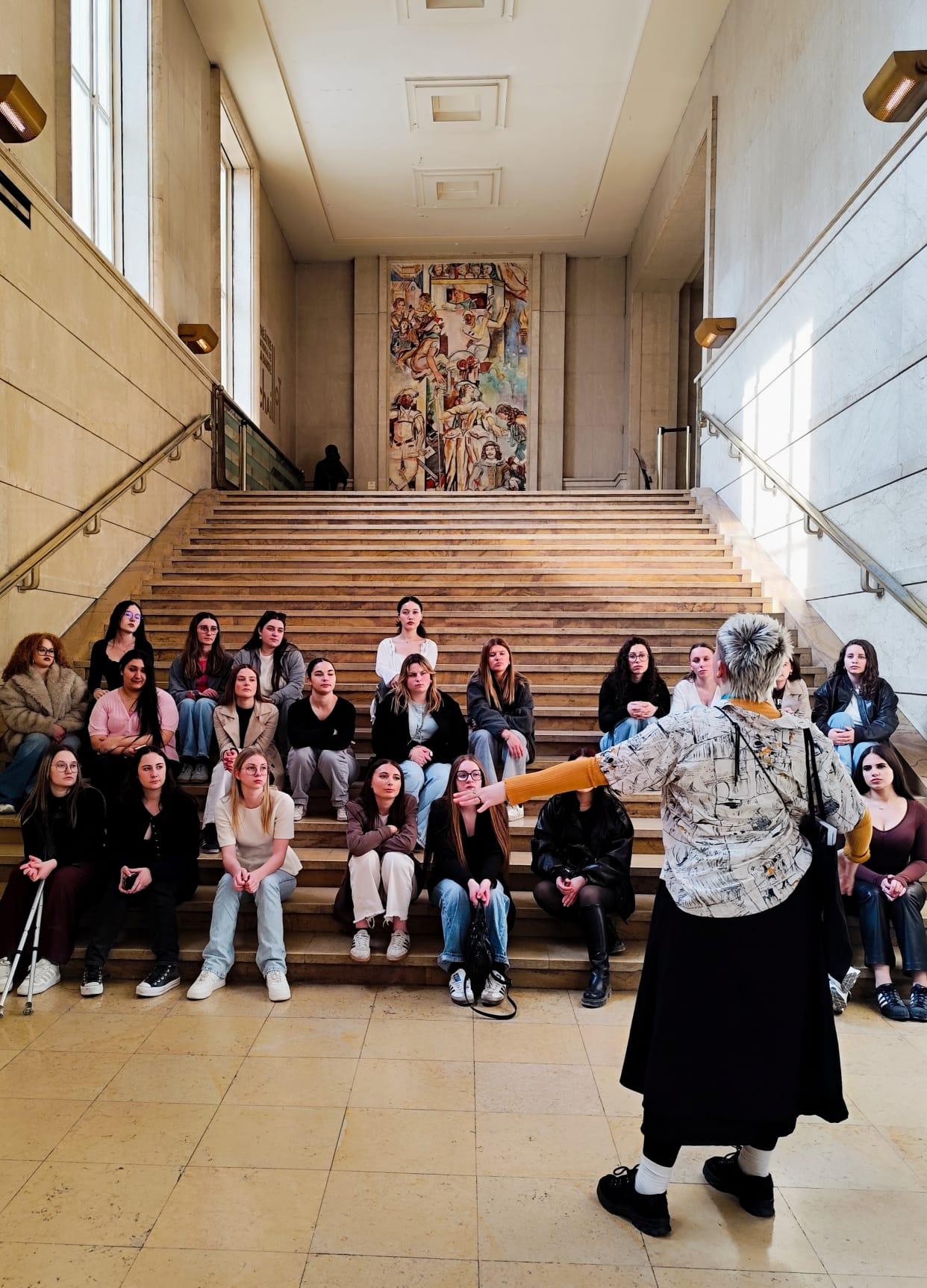 Le groupe est assis dans le grand escalier, face à la médiatrice. Au fond la fresque monumentale "Comédie".