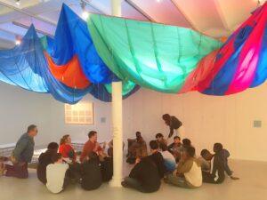 Les élèves sont assis sous une toile de parachute tendue au plafond. Ce toit textile coloré est pendu assez lâche et les abrite de la lumière. 